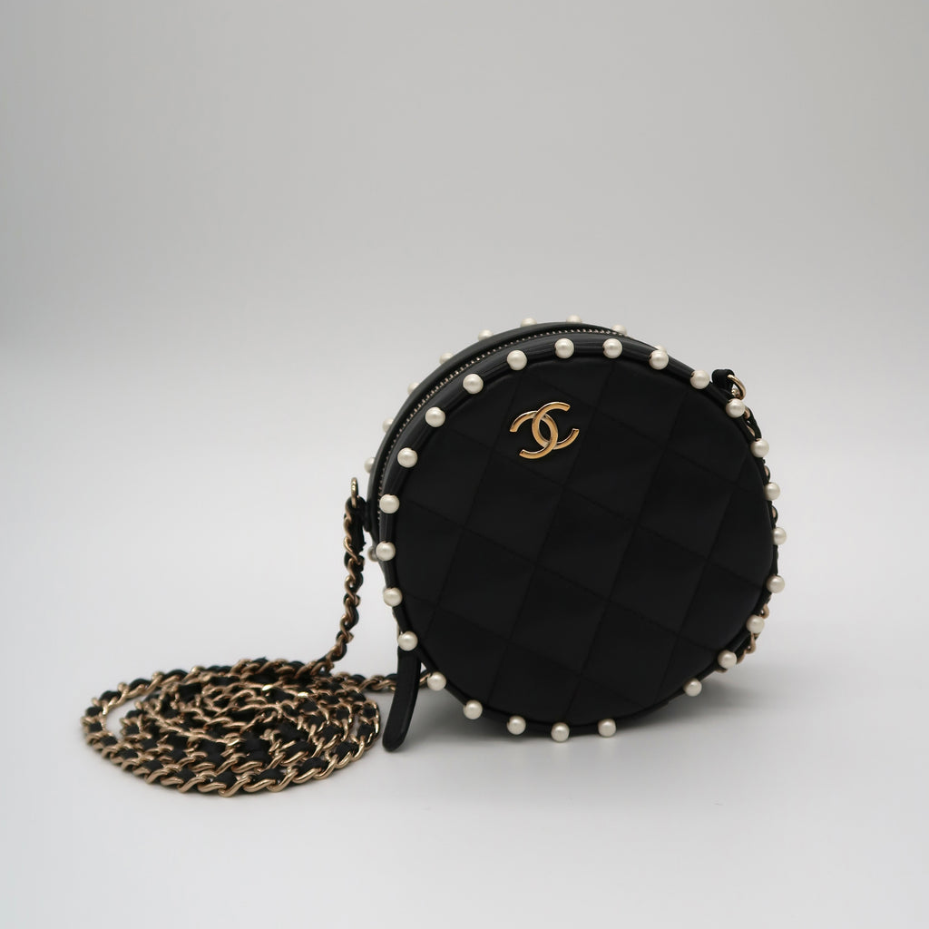 A black quilted Chanel round crossbody bag with a gold interlocking CC logo, adorned with white pearls around the edge, shown with its gold and black chain strap on a light gray background.