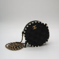 A black quilted Chanel round crossbody bag with a gold interlocking CC logo, adorned with white pearls around the edge, shown with its gold and black chain strap on a light gray background.