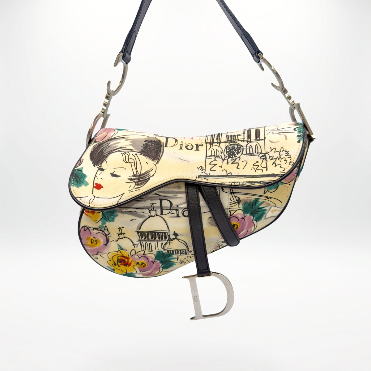 A Dior Saddle bag in cream canvas with a colorful illustration of a woman's face, floral motifs, and architectural sketches, featuring black leather straps and a silver-tone 'D' charm.