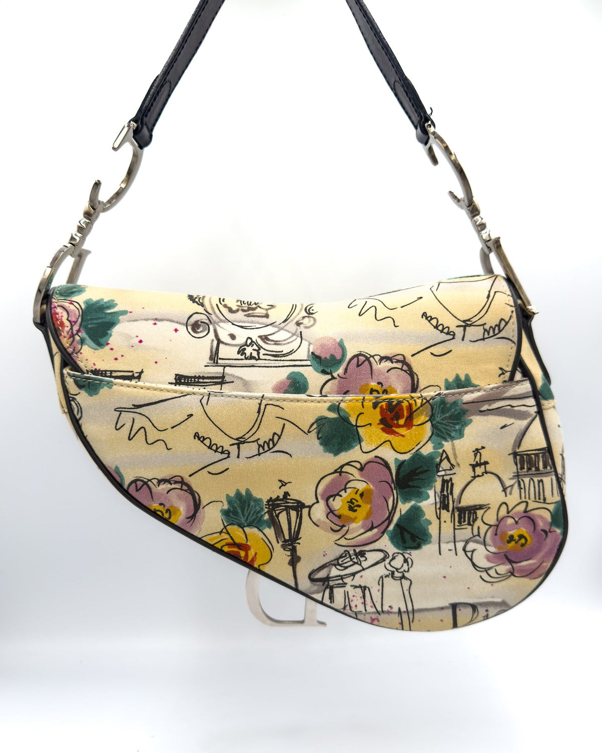 A vintage-style crescent shoulder bag with a cream-colored background, decorated with a hand-painted floral pattern in pink, yellow, and green, along with black sketch illustrations. It features a black leather strap and silver-tone hardware.