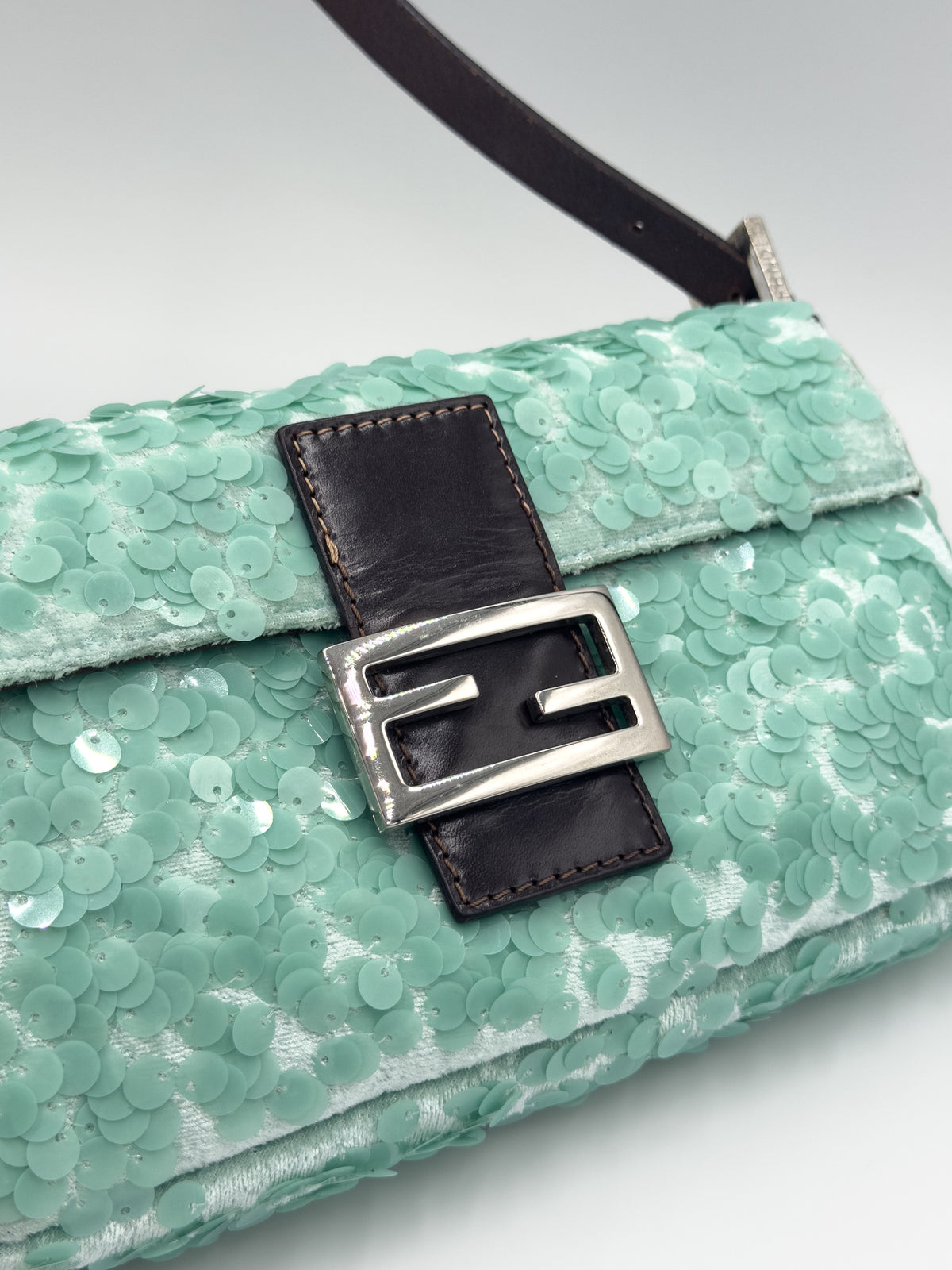 A close-up of a mint green sequined handbag with a dark brown leather strap detail and a silver-tone buckle featuring an interlocking F design, indicating the brand Fendi.