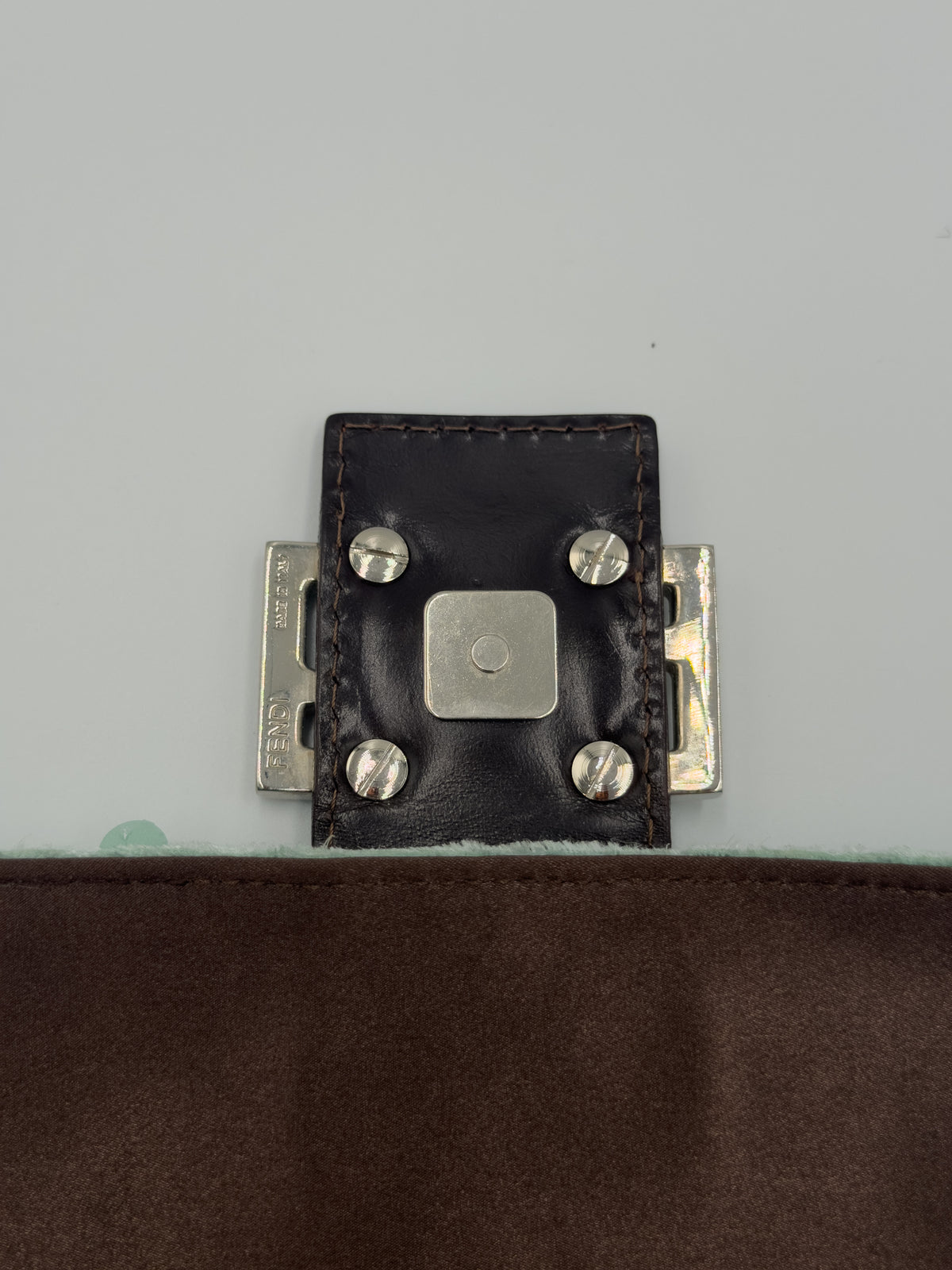 Close-up of a dark brown leather strap closure with silver-tone metal rivets and a central square metal plate, shown against a light gray background with a brown fabric surface below.