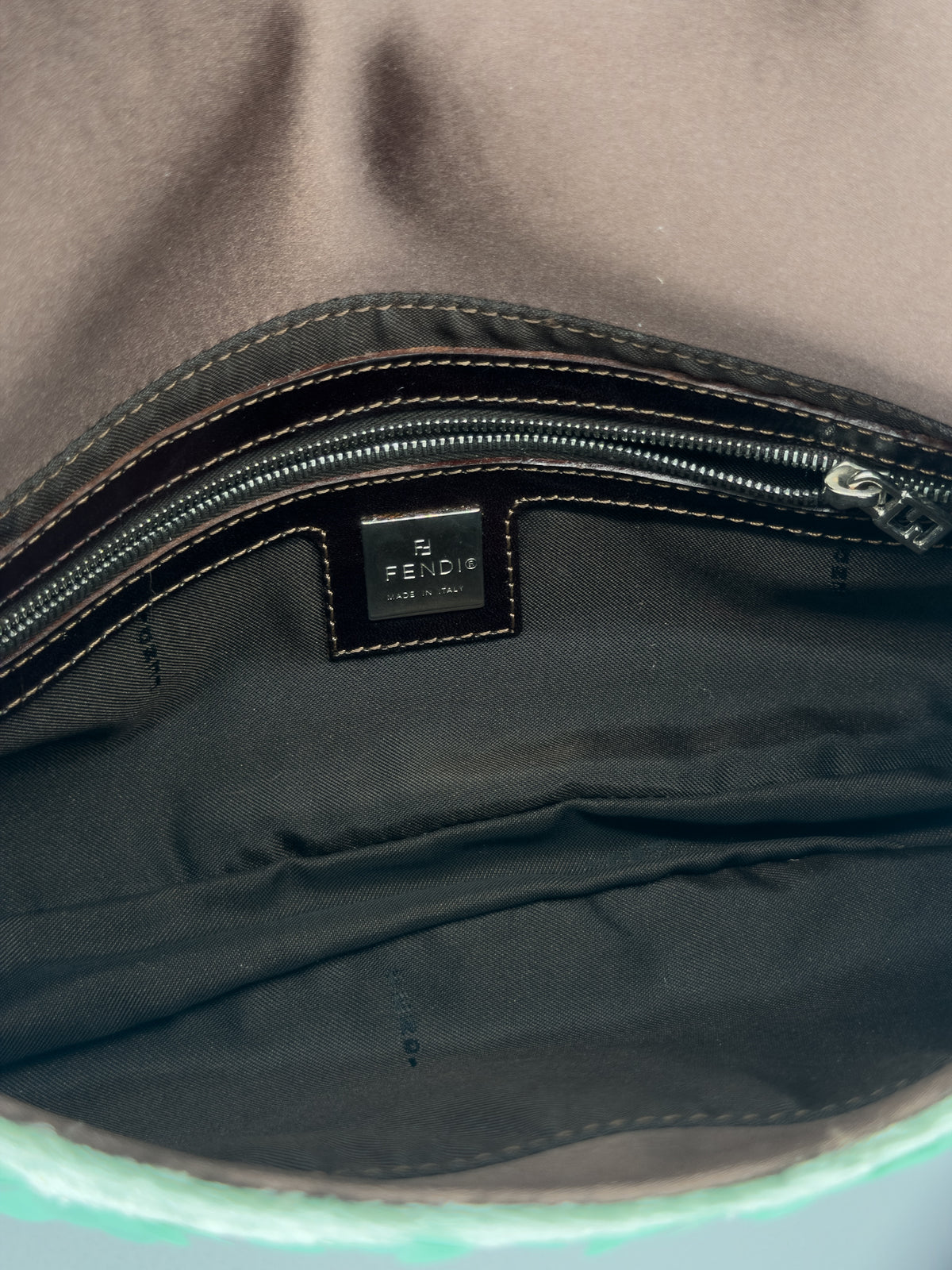 Close-up of the interior of a Fendi bag, showing a black leather brand label stitched onto a black nylon lining, with a brown fabric exterior visible above the zipper.