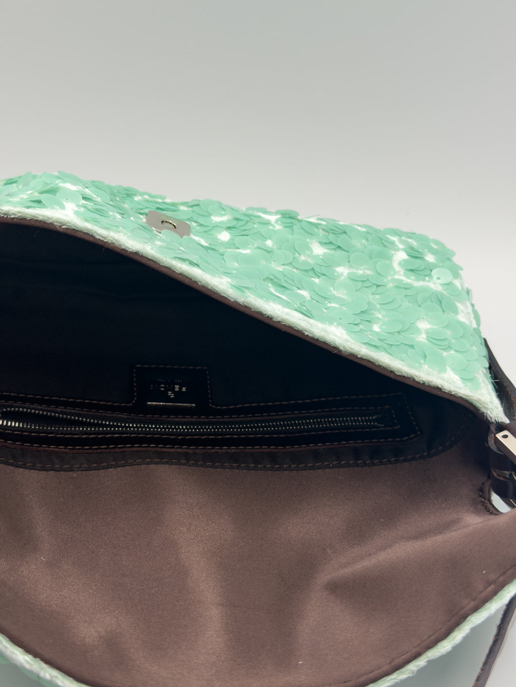 Close-up of an open handbag showing a mint green textured exterior flap and a brown satin-lined interior with a zippered pocket.