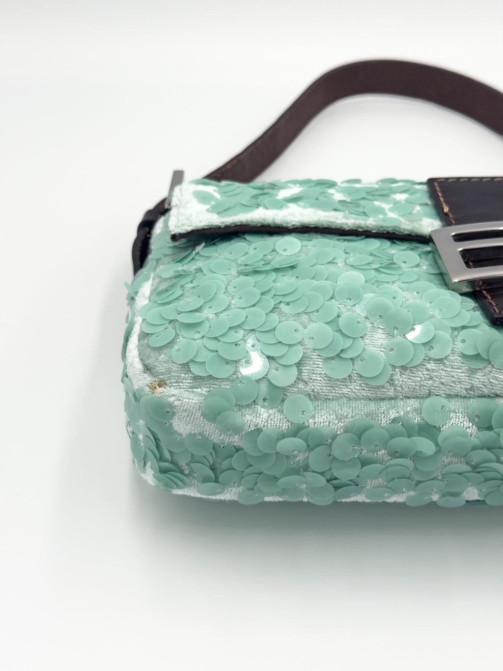 A close-up of a mint green sequined handbag with a dark brown strap and a silver-tone buckle, set against a plain white background.